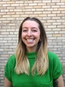 Image of Jordan Swartz, a white woman with long brown/blonde hair. She is smiling and wearing a short-sleeved green sweater. She is standing in front of a tan brick wall. 