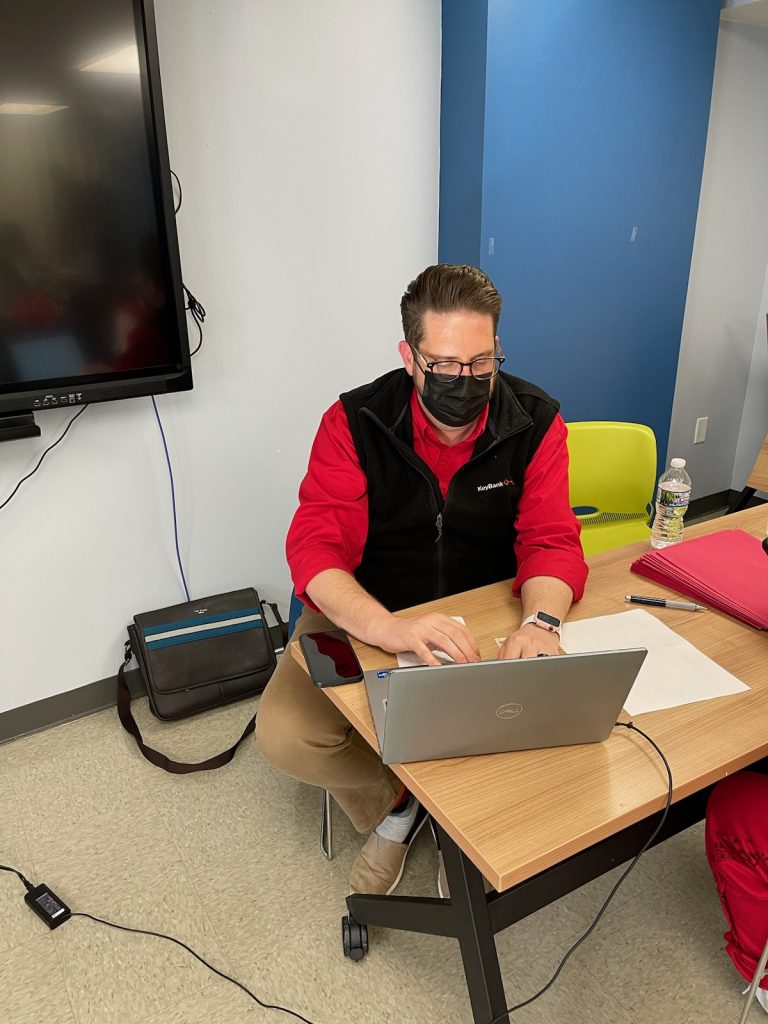 Image of a white man wearing a bright red button down shirt and black lightweight zip-up vest. He also wears a mask and glasses. He sits behind a silver laptop, focusing on it.