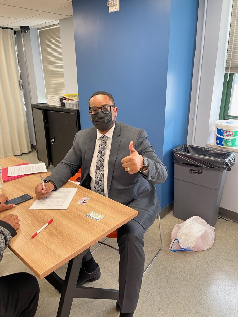 Image of a Black man with rectangular glasses and wearing a gray suit. He is sitting across a light, wooden table from an account opener. He has paperwork in front of him, and looks at the camera with a "thumbs up." He also wears a mask.